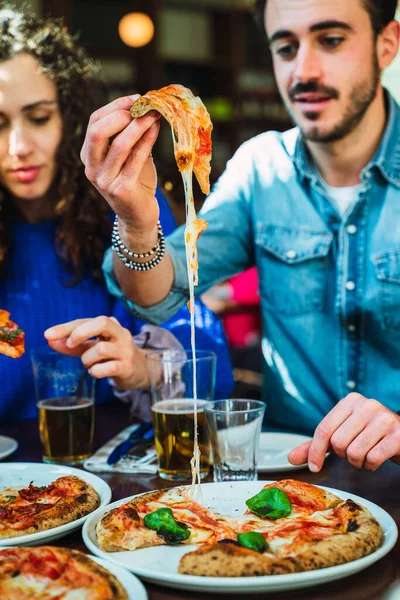 Guy taking a pizza slice out from the plate. The cheese stretching and looking appetizing. Friends enjoying cheese pizza with crispy crust. Beer glasses on the table.