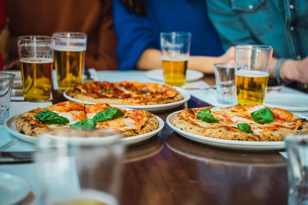 Pizzas and beer in glasses on a wooden table. Friends having pizza party after work. Closeup shot of three pizzas on white plates. Lunch at work. Sharing meal concept.