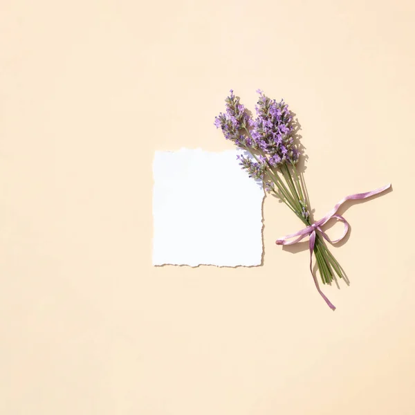 A bouquet of lavender flowers with a purple ribbon and white paper with torn edges on a pastel beige background. Minimal concept. Flat lay.