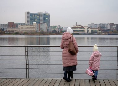 Anne ve kızı ördeklerle göle bakıyor. Rusya 'da bulutlu, sonbahar günü. Soğuk hava. Şehir manzarası, sokak fotoğrafı.