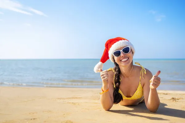 Noel Baba şapkalı ve güneş gözlüklü mutlu kadın kumsalda yatıyor. — Stok fotoğraf