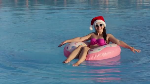Glimlachende jonge vrouw in santa hoed en zonnebril ontspannen op opblaasbare ring in het zwembad — Stockvideo