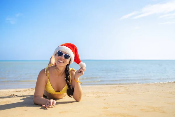 Счастливая женщина в шляпе санта и солнцезащитных очках, лежащая на песчаном пляже — стоковое фото