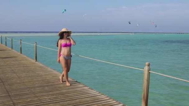 Wanita muda bahagia berkacamata hitam dan topi jerami berjalan di ponton kayu — Stok Video