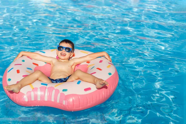 Güneş gözlüklü gülümseyen küçük çocuk yüzme havuzunda şişme halkada dinleniyor.