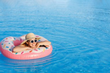 Hasır şapkalı ve güneş gözlüklü genç bir kadın yüzme havuzunda şişme yüzükte dinleniyor.