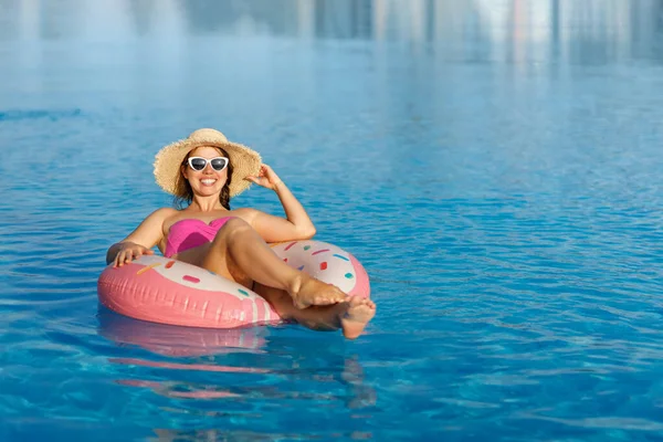 Sorridente jovem mulher em chapéu de palha e óculos de sol relaxante no anel inflável na piscina — Fotografia de Stock