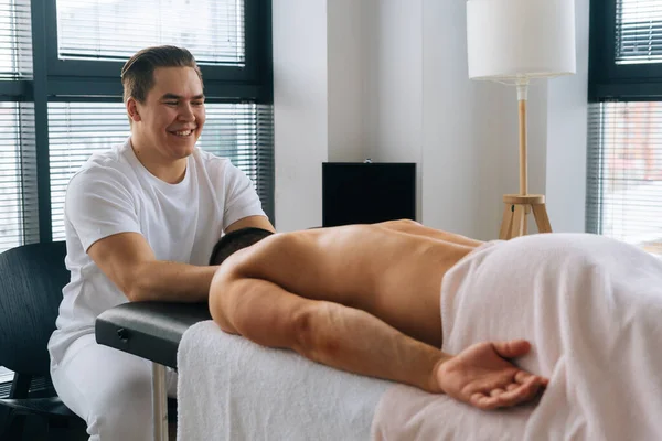 Side view of cheerful professional male masseur with strong hands massaging shoulder of muscular sports man lying on stomach at massage table
