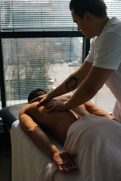 Vertical back view of professional male masseur with strong tattooed hands massaging back and shoulders of muscular sports man lying on stomach