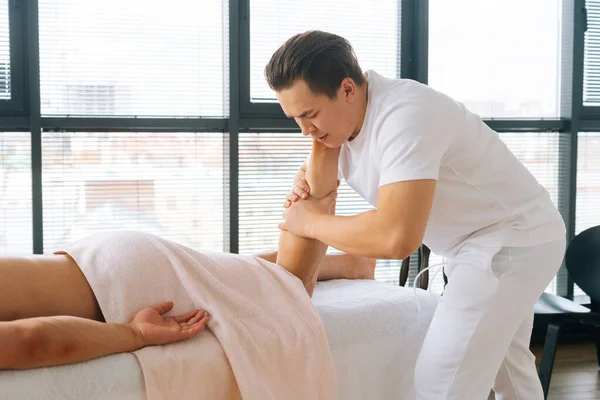 Cropped side view of professional male masseur with strong hands massaging calf muscles on lower leg of muscular athlete after physical sports workout