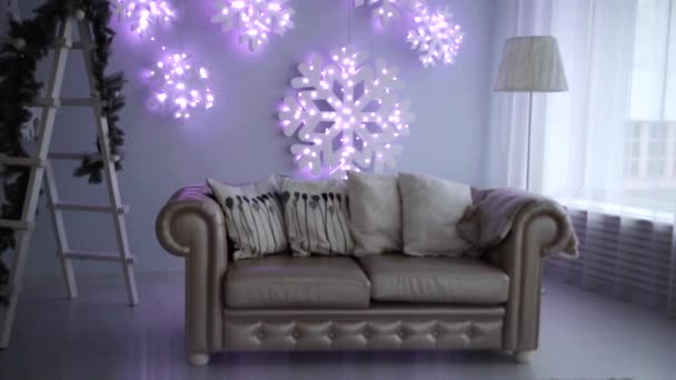 Decoração de Ano Novo da sala de estar. Há um sofá com um cobertor e travesseiros na sala, uma escada com uma decoração de galhos de árvore de Natal pendurados na parede brilhante flocos de neve — Vídeo de Stock