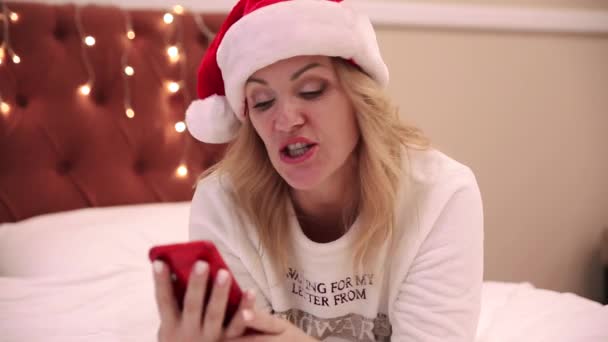 산타 클라우스 모자를 쓴 한 여성 이 침대에 누워 휴대폰을 보고 메시지를 인쇄하고 축하의 메시지를 보냅니다. 크리스마스와 새해 분위기 — 비디오