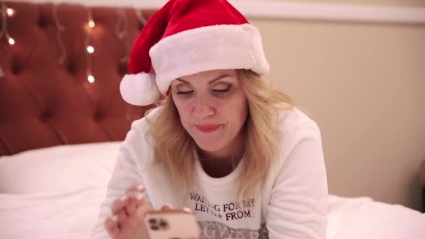 Kobieta w czapce Świętego Mikołaja leży na łóżku i patrzy na telefon komórkowy, drukuje wiadomości i gratuluje. Boże Narodzenie i Nowy Rok atmosfera — Wideo stockowe