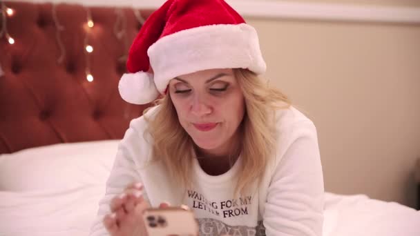 Una donna con un cappello da Babbo Natale si trova sul letto e guarda un telefono cellulare, stampa messaggi e congratulazioni. Natale e Capodanno atmosfera — Video Stock