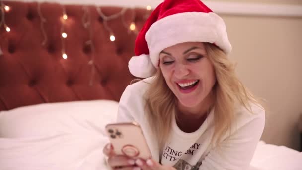 Женщина в белом свитере и шляпе Санта Клауса лежит на кровати и смотрит на мобильный телефон и поет песни. Новогодняя и рождественская атмосфера — стоковое видео
