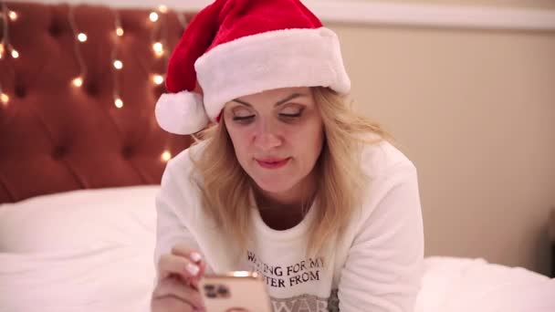 Une femme au chapeau de Père Noël est allongée sur le lit et regarde un téléphone portable, imprime des messages et des félicitations. Ambiance Noël et Nouvel An — Video