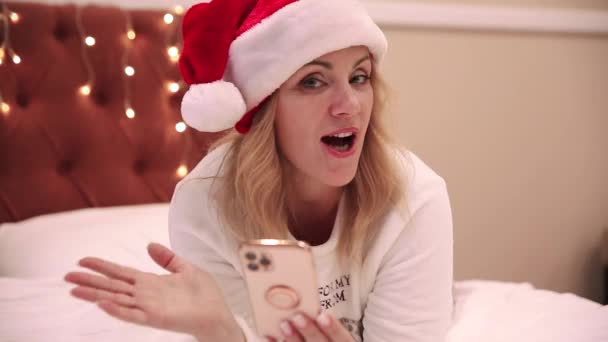 Egy nő fehér pulóverben és télapó sapkában fekszik az ágyon, ránéz egy mobiltelefonra és dalokat énekel. Karácsony és újév hangulata — Stock videók