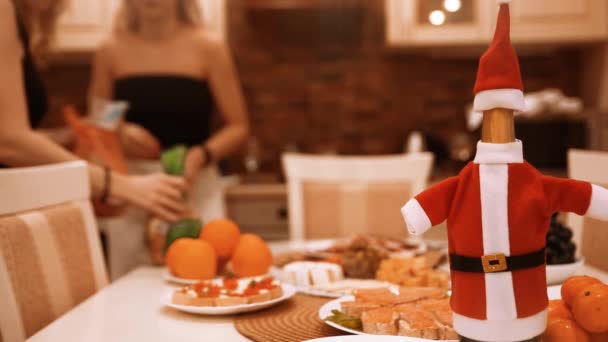 Друзья-женщины собираются вместе и готовят рождественский или новогодний ужин, накрывают на стол, накрывают на посуду. Сосредоточьтесь на бутылке вина в костюме Санта Клауса. — стоковое видео