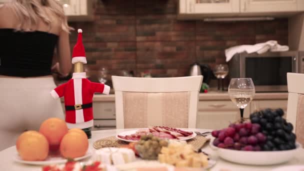 Freundinnen treffen sich und bereiten Weihnachts- oder Neujahrsessen zu, decken den Tisch, decken das Geschirr. Fokus auf eine Flasche Wein im Weihnachtsmann-Kostüm. — Stockvideo