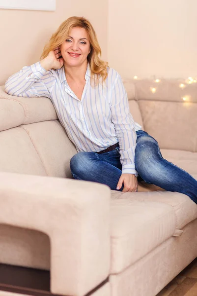 一个穿着衬衫和牛仔裤的漂亮女人正坐在家里的沙发上 看着相机笑着 圣诞及新年气氛 — 图库照片