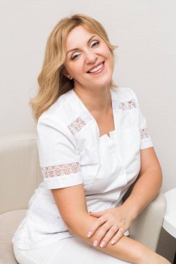 Beyaz üniformalı güzel bir güzellik doktoru kameraya bakıp ofisinde bir sandalyede otururken poz veriyor. Dikey fotoğraf