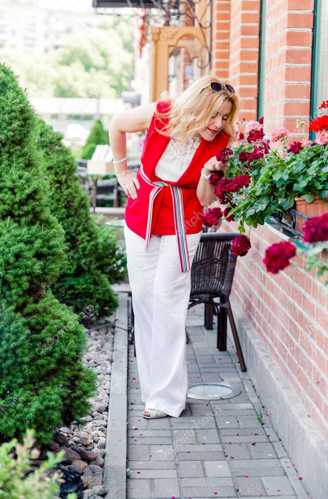 Beautiful mature woman in a white and red suit poses against a brick wall and sniffs a flower. Woman happy life concept after 40 years
