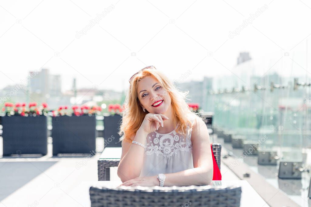 A beautiful mature woman in a white blouse sits on a summer terrace and enjoys the weather. Woman happy life concept after 40 years