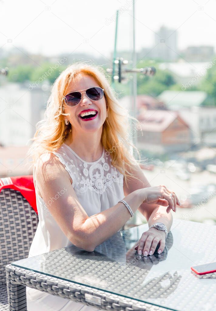 A beautiful mature woman in a white blouse sits on a summer terrace and enjoys the weather. Woman happy life concept after 40 years