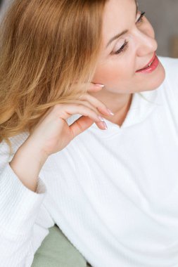 Sevimli orta yaşlı sarışın bir kadın. Işıl ışıl gülümseyen bir kadın. Evdeki kanepeye yakın oturuyor.