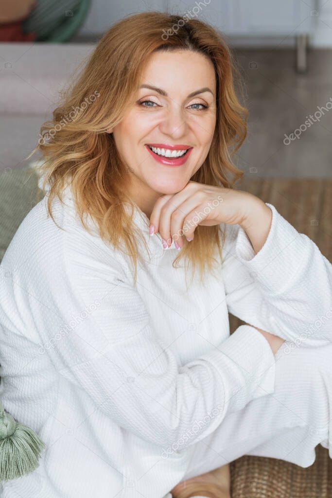 Lovely middle-aged blond woman with a beaming smile sitting next to sofa at home looking at the camera. Photo
