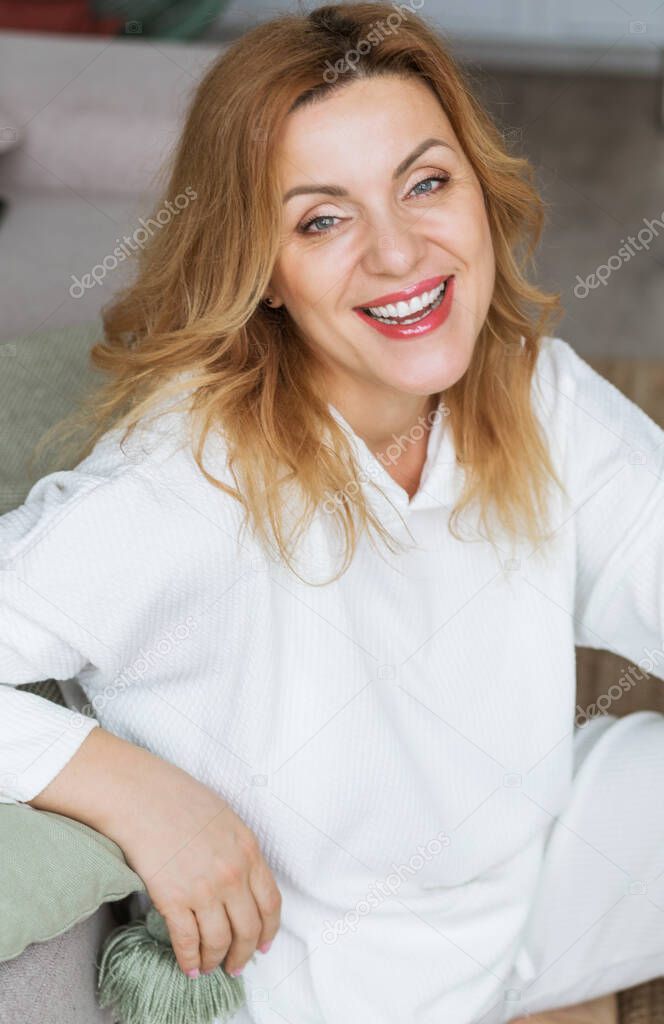 Lovely middle-aged blond woman with a beaming smile sitting next to sofa at home looking at the camera. Photo