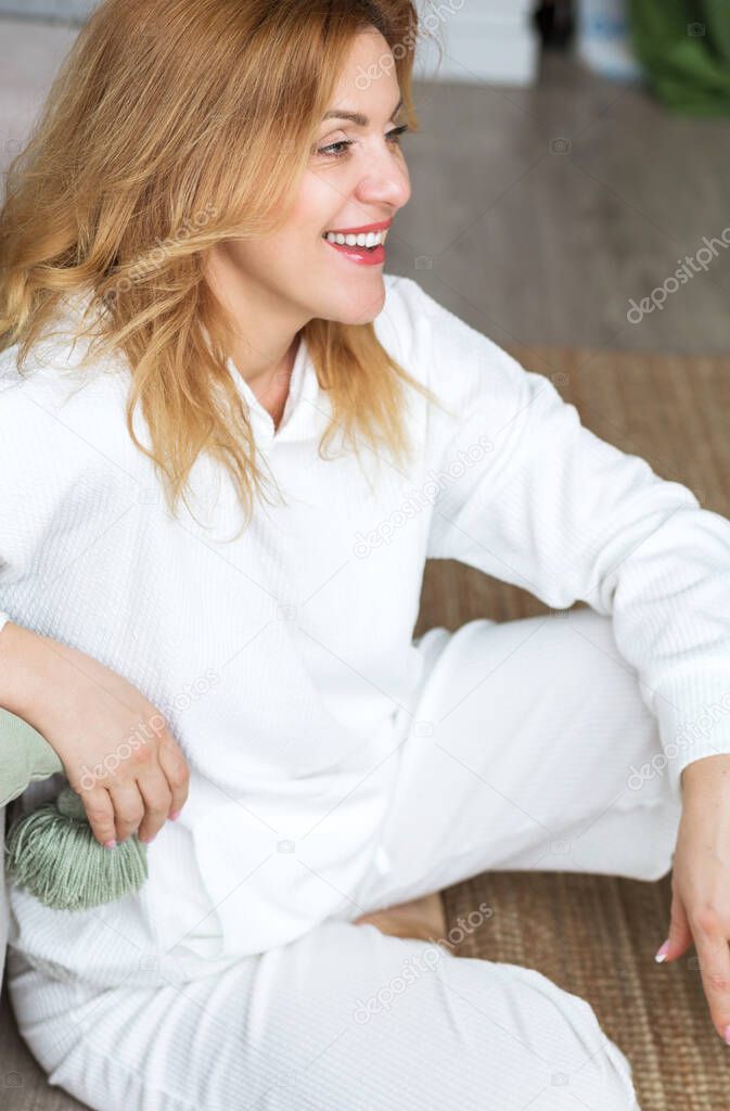Lovely middle-aged blond woman with a beaming smile sitting on the floor close to the sofa at home looking somewhere