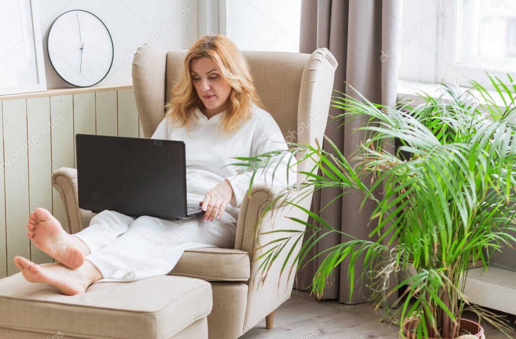 Beautiful mature smiling woman working on laptop and sitting in a big comfortable chair at home. Female entrepreuneur and freelance concept