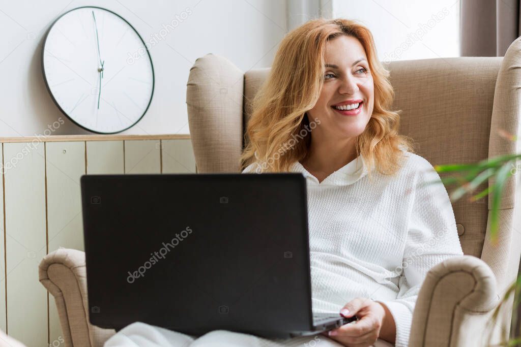 Beautiful mature smiling woman working on laptop and sitting in a big comfortable chair at home. Female entrepreuneur and freelance concept
