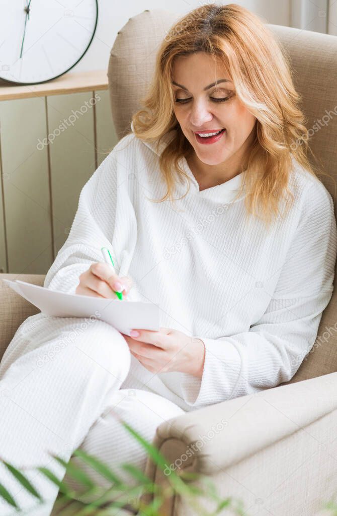 A woman in white clothes sits in a home chair and writes her dreams on paper. Desire visualization concept