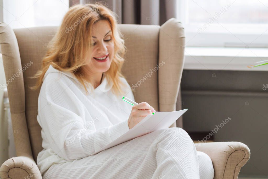 A woman in white clothes sits in a home chair and writes her dreams on paper. Desire visualization concept