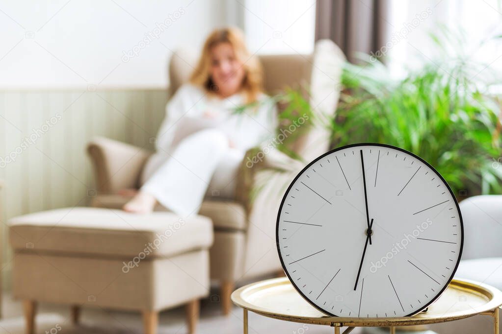 In the foreground there is a clock on a table, in the background a woman is sitting. The concept of unhurried life and relaxation at home