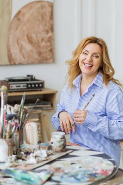 Olgun bir kadın stüdyoda tuvale fırça sürüyor ve kameraya bakıyor..