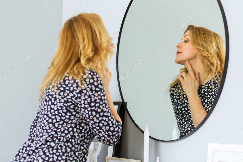 A beautiful mature woman in a fashionable dress stands in front of a mirror, touches her skin and assesses its condition. Woman happy life concept after 40 years
