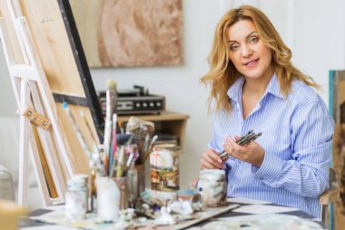 Olgun bir kadın stüdyoda tuvale fırça sürüyor ve kameraya bakıyor..