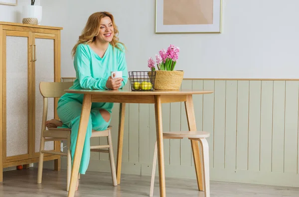 A beautiful woman in a green home suit sits in the kitchen at the table and drinks tea