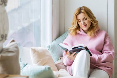 Pencere kenarında oturan ve kitap okuyan bir kadın. Sosyal medya detoksu. Yavaş yaşam tarzı. Edebiyattan hoşlanan mutlu kadın. Sakin kitap kurdu, bütün günü aletlerle geçiriyor.