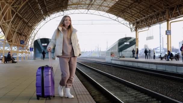 Młoda kobieta w ciepłym garniturze i kurtce z walizką czeka na pociąg na peronie dworca kolejowego. — Wideo stockowe