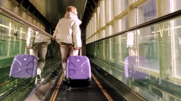 Mujer joven en la pasarela en movimiento en el aeropuerto con una maleta púrpura. — Vídeos de Stock