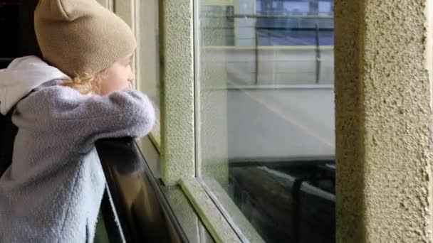 En liten flicka åker rulltrappa och tittar ut genom fönstret på järnvägsspåren och tågen — Stockvideo