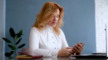 Gülümseyen beyaz bir kadın elinde akıllı telefonu tutuyor.