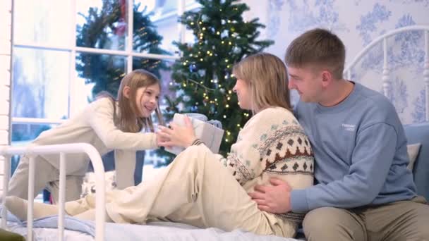 Pappa och mamma sitter på sängen, dottern ger mamma en julklapp — Stockvideo