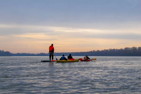 Friends in kayaks and on stand up paddle board (paddleboard, SUP) rowing with a paddle and in the winter Danube river at calm cold winter evening