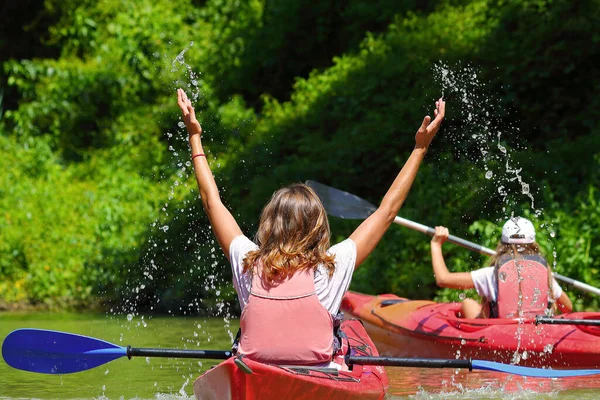 Kırmızı kanoyla su sıçratan mutlu sarışın kadın. Elleriyle havaya fırlatırken su kahkaha atıyor. Yaz aylarında Tuna Nehri kıyısında yeşil ağaçlarla kano yaparken. Eğlence konsepti. Arka plan.