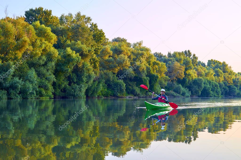 Woman paddle green kayak on Danube river against the background of green spring trees at sunrise. Concept for adventure, travel, action, lifestyle.